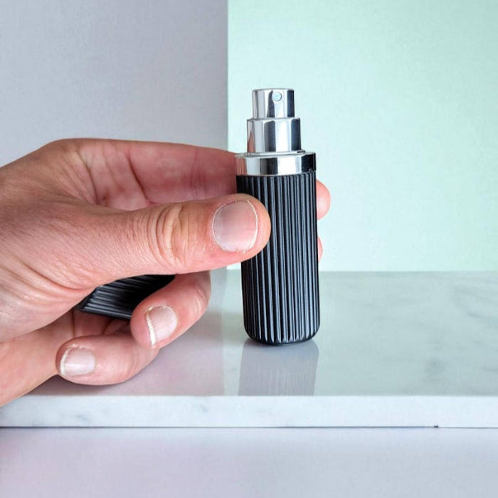 Hand holding a black and silver cylindrical perfume atomizer against a light background